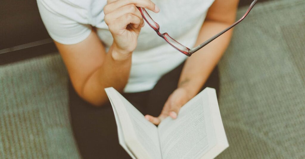 Young woman sitting indoors, reading a book while holding eyeglasses. A moment of focus and reflection.