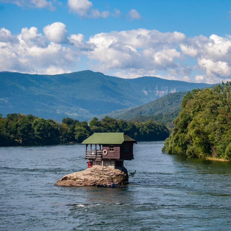 Drvena kućica na maloj steni u sred Drina reke, okružena prirodom, zelenilom i planinama u daljini