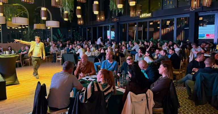 Grupe ljudi okupljene oko stolova u velikom prostoru u opuštenoj atmosferi , koji prate voditelja